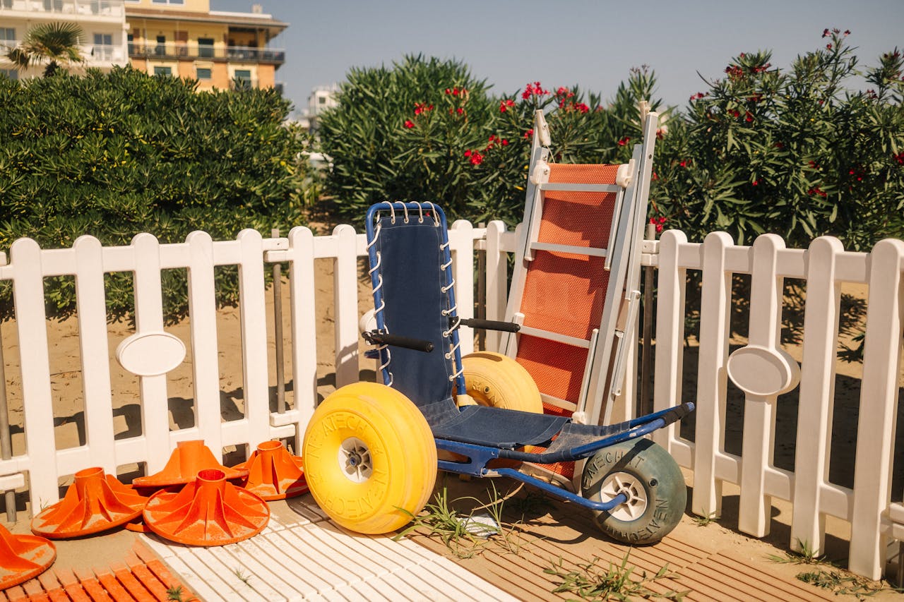 services-01 Beach wheelchair next to lifeguard equipment by a white fence on a sunny day.